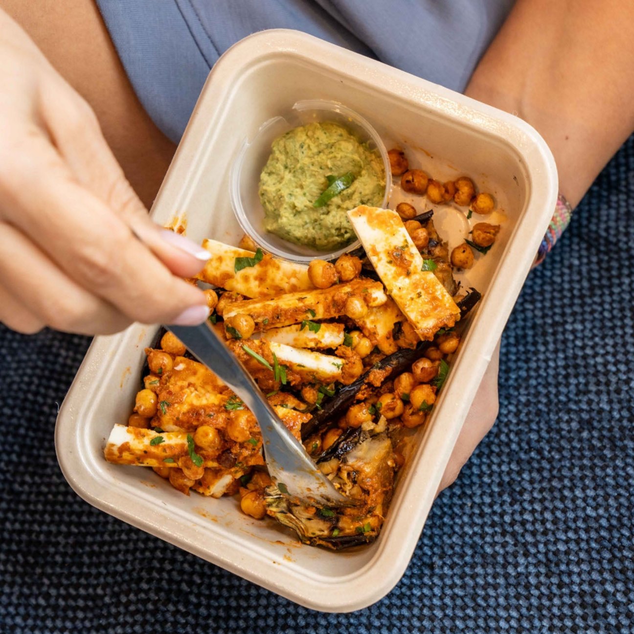 A close-up of a meal in a biodegradable container, featuring grilled paneer slices, roasted chickpeas, and charred vegetables, garnished with fresh herbs. A small portion of green chutney or dip is served in a separate container. The meal is being enjoyed with a fork, held by a hand, while resting on a textured blue surface