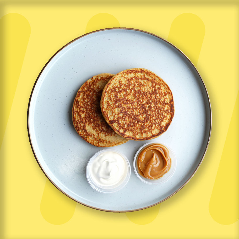 A plate featuring two golden-brown pancakes, served with small portions of creamy yogurt and smooth peanut butter in separate containers. The presentation is minimalist, with the pancakes placed on a light ceramic plate with a dark rim, set against a bright yellow background that complements the dish's warm tones.