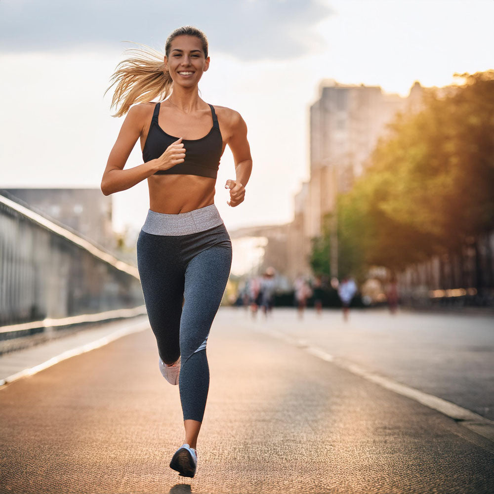 A fit woman jogging on a sunlit city street, wearing a black sports bra and gray leggings, with her hair in a ponytail and a bright smile.