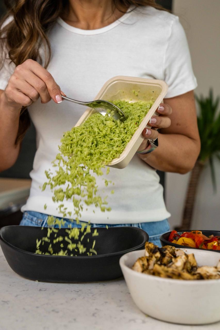 Close-up of a person wearing a white T-shirt and jeans, pouring vibrant green shredded vegetables or grains into a black bowl using a spoon. Nearby are two additional bowls, one with grilled chicken pieces and the other with roasted colorful bell peppers, all placed on a light-colored countertop