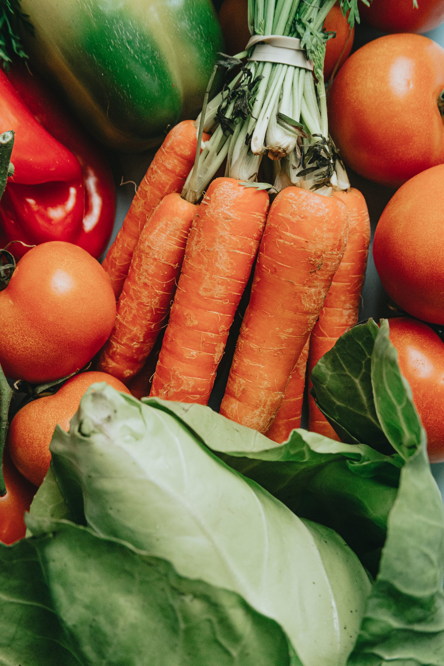 A colorful assortment of fresh vegetables, including bell peppers, carrots, tomatoes, and leafy greens, arranged on a rustic wooden surface.