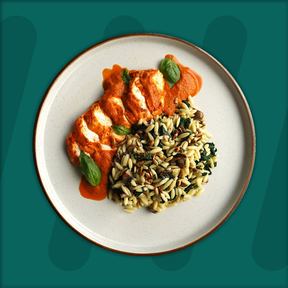 A plate featuring sliced chicken breast drizzled with a rich tomato-based sauce and garnished with fresh basil, served alongside a portion of orzo pasta mixed with sautéed spinach, mushrooms, and toasted pine nuts, set against a green background.