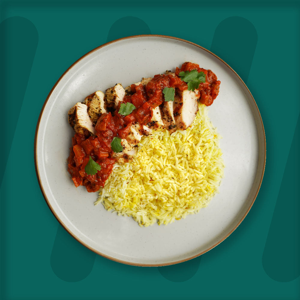 prepped meal, A vibrant plate featuring grilled chicken slices topped with a flavorful tomato-based curry sauce, garnished with fresh cilantro leaves. Served alongside is a portion of fluffy yellow rice, perfectly complementing the dish. The meal is presented on a light ceramic plate with a dark rim, set against a rich green background to highlight its appetizing colors