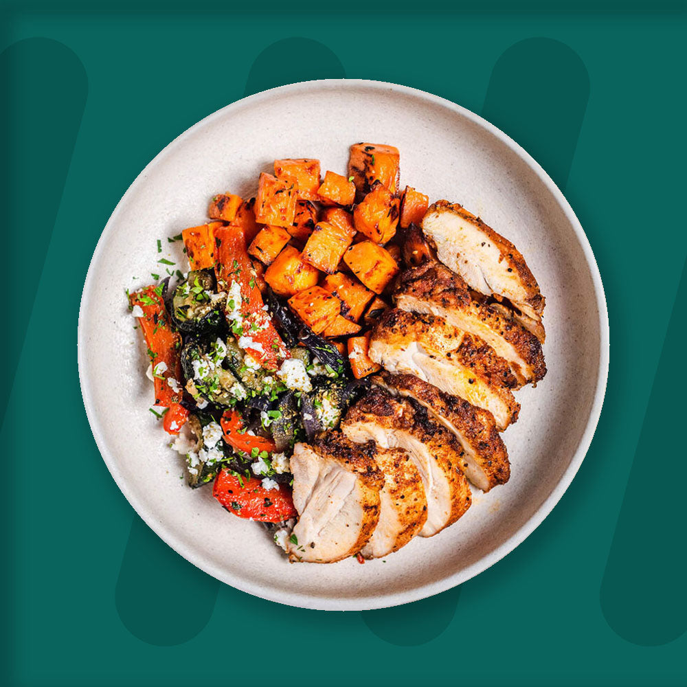 A hearty and colorful plate featuring seasoned grilled chicken slices, paired with roasted sweet potato cubes and a medley of roasted vegetables, including zucchini, eggplant, and bell peppers, topped with crumbled feta cheese and fresh herbs. The dish is served in a simple ceramic bowl, set against a rich green background that accentuates the vibrant and inviting colors of the meal