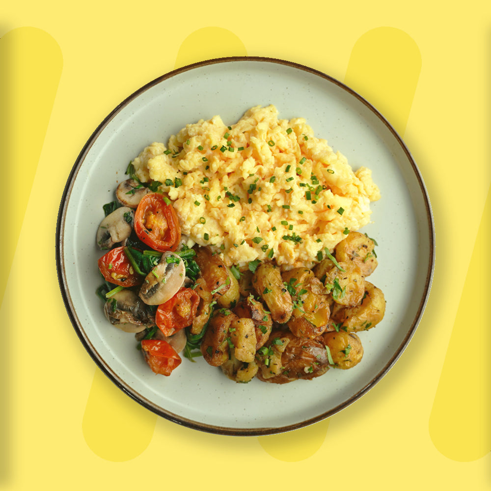 A beautifully plated breakfast featuring creamy scrambled eggs topped with chopped chives, alongside roasted baby potatoes with herbs and a medley of sautéed spinach, mushrooms, and cherry tomatoes. The dish is served on a ceramic plate with a dark rim, set against a bright yellow background that highlights the fresh and hearty meal