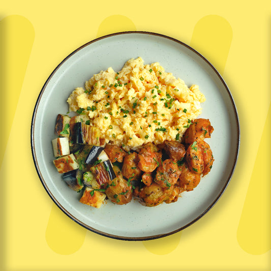 A vibrant plate featuring fluffy scrambled eggs garnished with fresh chives, roasted baby potatoes tossed in harissa spices, and grilled cubes of eggplant with herbs. The meal is elegantly presented on a light ceramic plate with a dark rim, set against a cheerful yellow background that enhances the dish's appetizing colors.