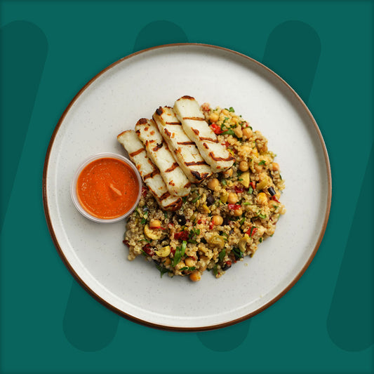 A plate featuring grilled slices of halloumi cheese served on a bed of quinoa salad mixed with chickpeas, diced vegetables, and fresh herbs, accompanied by a small bowl of vibrant red pepper sauce, set against a green background.