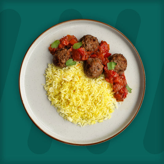 A plate of seasoned meatballs topped with a vibrant tomato-based sauce, served alongside fluffy yellow rice, garnished with fresh cilantro, set against a teal background