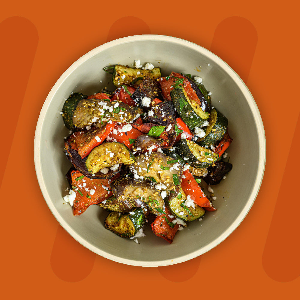 A bowl of roasted mixed vegetables including zucchini, bell peppers, and eggplant, garnished with crumbled feta cheese and fresh herbs, set against an orange background