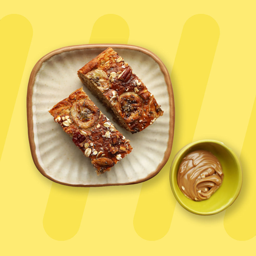 A beautifully presented plate featuring two slices of baked oat bars topped with banana slices, pecans, and oats for a wholesome and textured finish. The bars are served on a beige ceramic plate with a rustic rim, accompanied by a small green bowl filled with creamy peanut butter. The vibrant yellow background adds a warm and inviting touch to the presentation