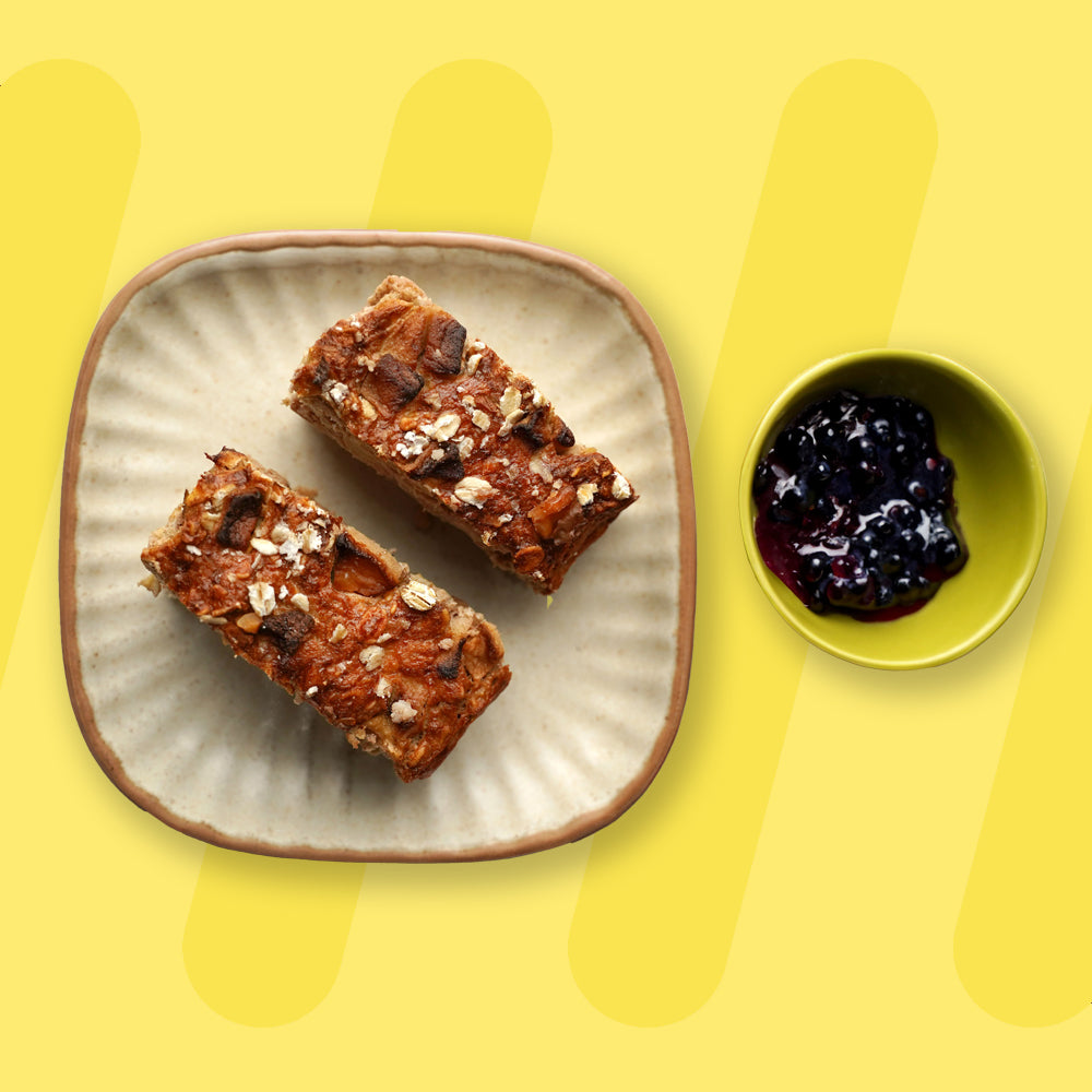 A minimalist presentation of two oat and nut bars, topped with rolled oats and roasted nuts, served on a textured beige ceramic plate. Accompanying the bars is a small vibrant green bowl filled with a rich blueberry compote, set against a bright yellow background for a cheerful and appetizing display