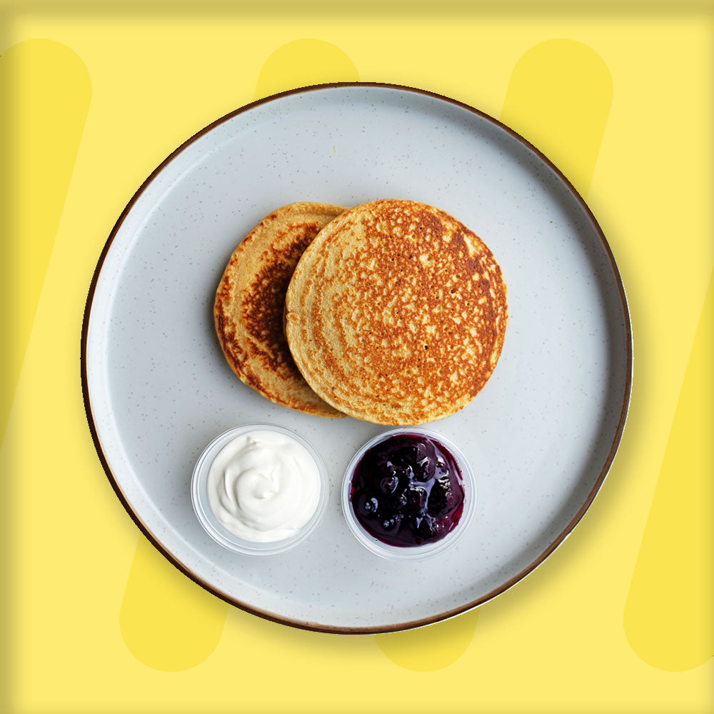 A plate featuring two perfectly golden pancakes, served with small portions of creamy yogurt and a rich blueberry compote in separate containers. The presentation is simple yet inviting, with the pancakes arranged on a light ceramic plate with a dark rim, set against a cheerful yellow background that highlights the dish's warmth and freshness